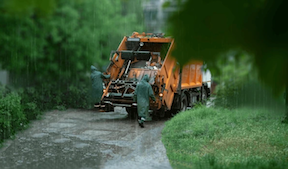 Collection truck driving in rain storm on narrow road with collectors on the back in raincoats